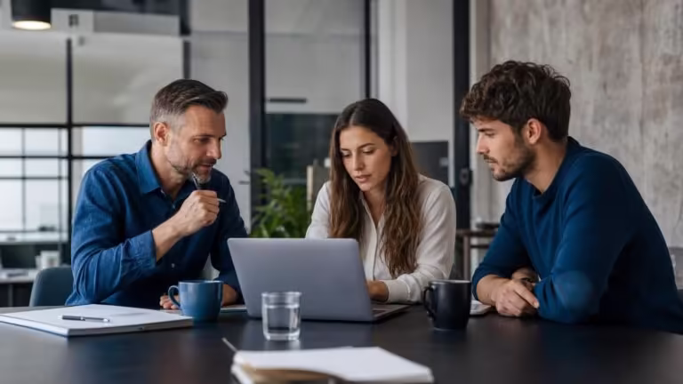 Three professionals are sitting at a table in an office, looking intently at a laptop screen during a collaborative meeting.